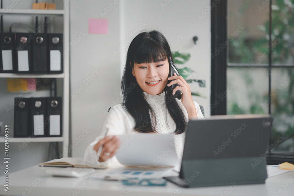 Businesswoman is multitasking at her desk, talking on the phone while reviewing paperwork Businesswoman is multitasking at her desk, talking on the phone while reviewing paperwork