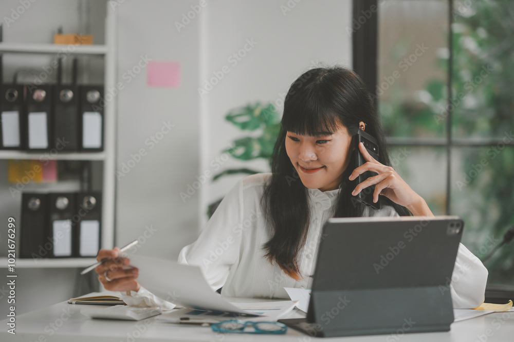 Asian accountant woman is working in her office, analyzing financial documents and talking on the phone Asian accountant woman is working in her office, analyzing financial documents and talking on the phone