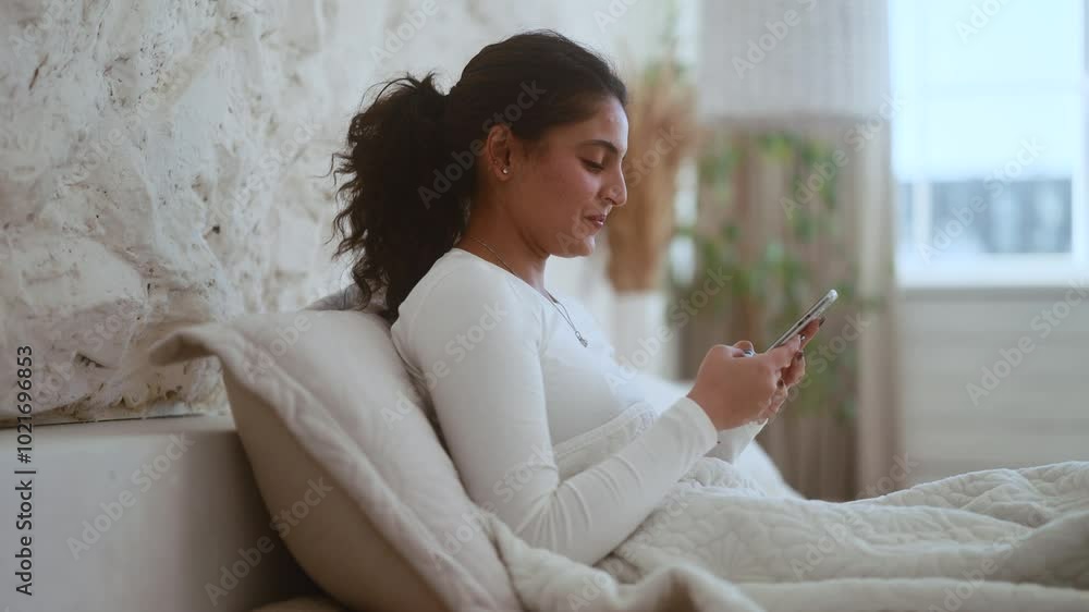 Charming smiling young indian woman hold smartphone scrolling app watching social media feed laying on bed indoors Happy female texting enjoying leisure time at cozy home Charming smiling young indian woman hold smartphone scrolling app watching social media feed laying on bed indoors Happy female texting enjoying leisure time at cozy home
