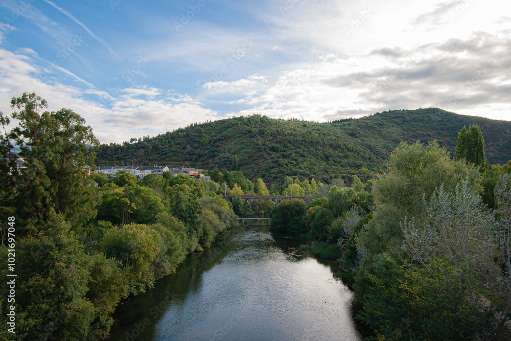 Fototapeta premium View of the Sil River as it passes through the city of Ponferrada under a pedestrian bridge and a railway bridge that gives access to the train station and the Pajariel mountain in the background