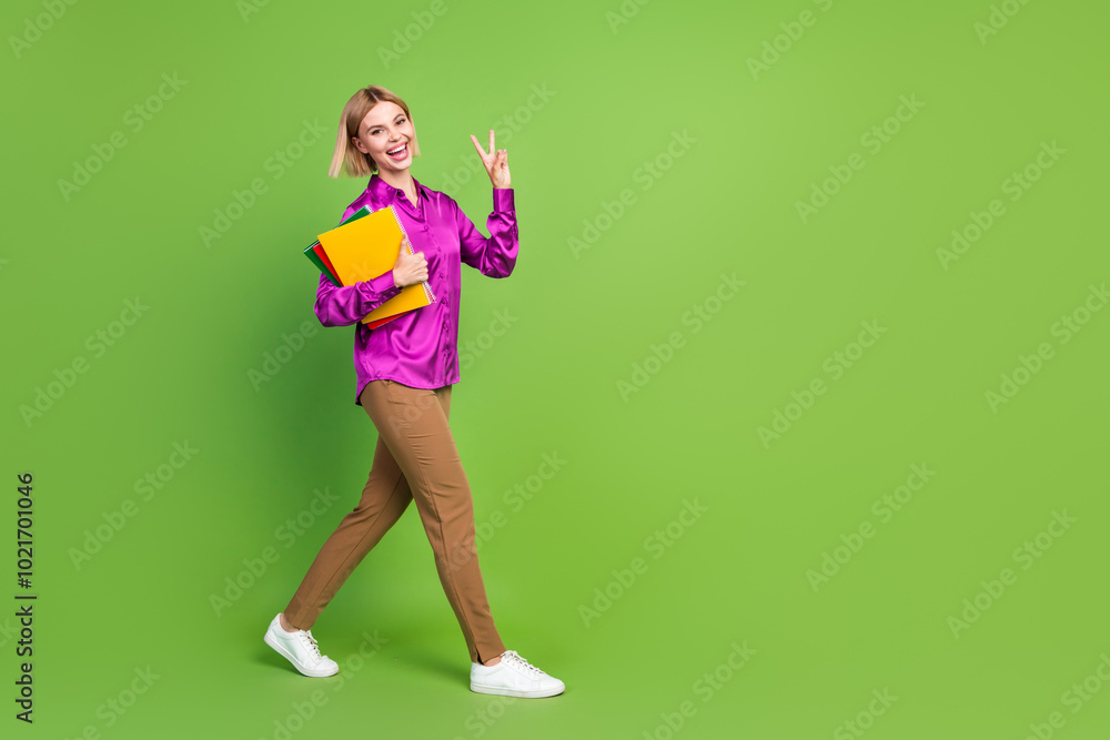 Full size photo of pretty young woman book walk empty space v-sign wear shirt isolated on green color background Full size photo of pretty young woman book walk empty space v-sign wear shirt isolated on green color background