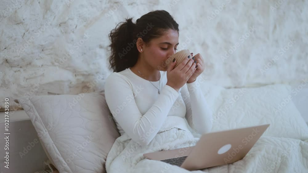 Pretty indian woman with cup of drink relaxing at home or working online browsing products in internet store reading news checking email or social media on laptop computer laying on bed in the morning Pretty indian woman with cup of drink relaxing at home or working online browsing products in internet store reading news checking email or social media on laptop computer laying on bed in the morning