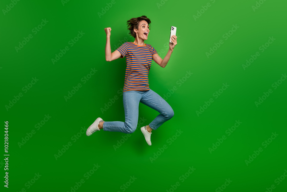 Full length profile portrait of excited energetic person jumo raise fist hold smart phone empty space isolated on green color background Full length profile portrait of excited energetic person jumo raise fist hold smart phone empty space isolated on green color background