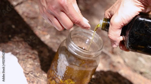 Herbalist pouring oil from a bottle into a jar filled with resins, showcasing the art of oily macerate preparation. Selective focus photo