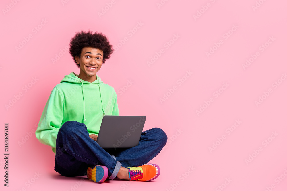 Full size photo of nice young man sit floor laptop wear green hoodie isolated on pink color background Full size photo of nice young man sit floor laptop wear green hoodie isolated on pink color background