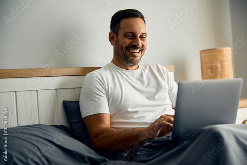 Happy man using laptop while relaxing in bed.