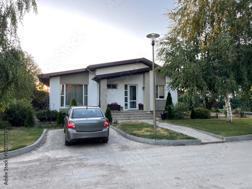 A quaint countryside house is set against a serene backdrop, featuring a parked car in the driveway. The evening light casts a warm glow on the well-maintained yard