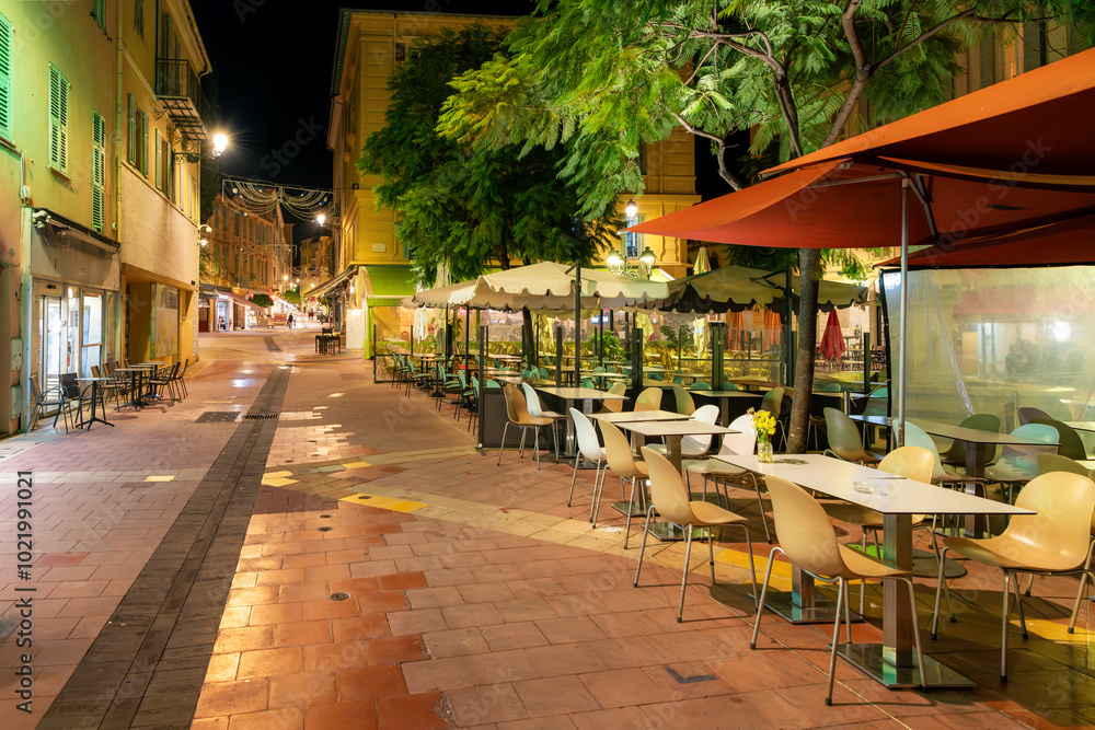 Naklejka premium Late night on the French Riviera in the illuminated historic old town district of Menton, France, with empty sidewalk cafes along the main street of shops.