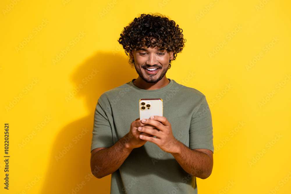 Photo of smart positive guy with wavy hairdo dressed khaki t-shirt look at smartphone typing message isolated on yellow color background