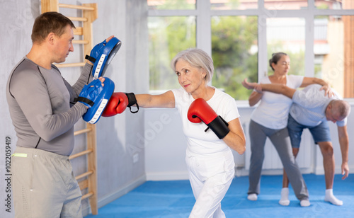 Senior lady and male couch are boxing in gym, man helps student to work out force of blow. Modern positive family chooses active hobby photo