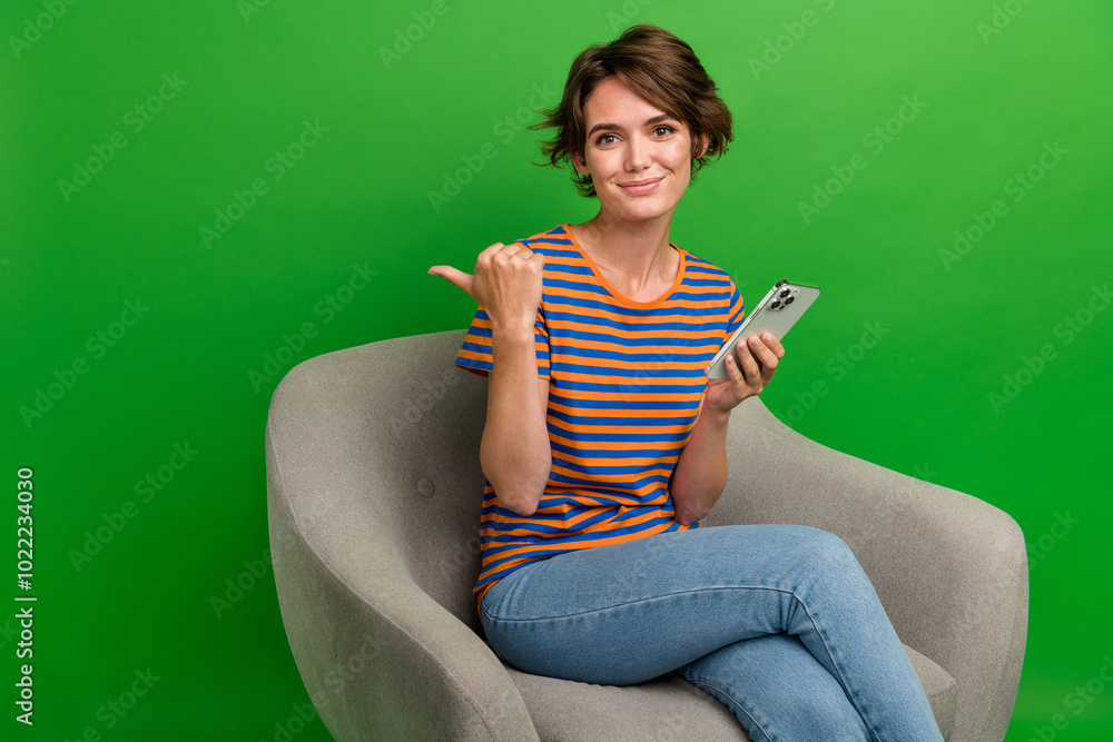 Portrait of charming person sit comfort chair hold smart phone direct finger empty space isolated on green color background Portrait of charming person sit comfort chair hold smart phone direct finger empty space isolated on green color background