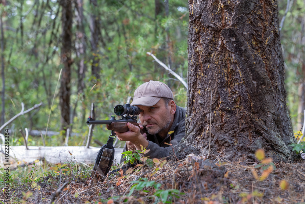 Hunter lying on the ground behind a tree, aiming his rifle at game while looking through the scope and ready to shoot. Hunter lying on the ground behind a tree, aiming his rifle at game while looking through the scope and ready to shoot.