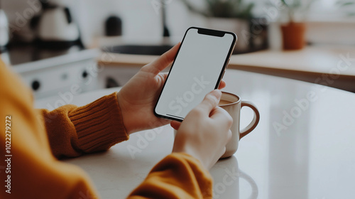 A high-quality image of a person holding a cup of coffee in one hand and using a smartphone with the other, sitting at a clean, minimalist kitchen table