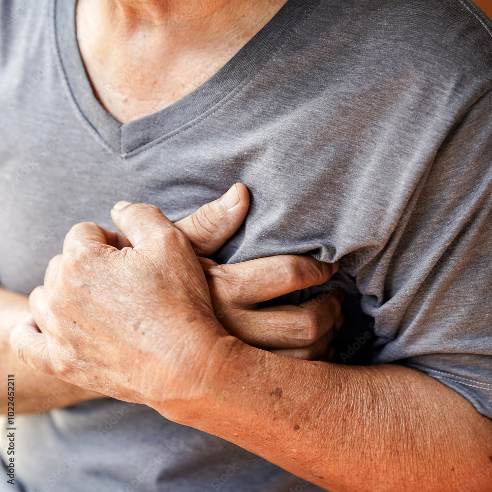 Fototapeta premium elderly person in gray shirt clutches their chest, possibly indicating discomfort or pain. image conveys sense of urgency and concern