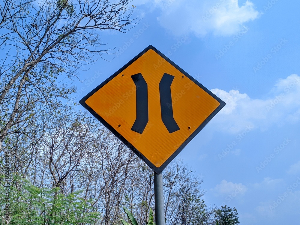 A yellow diamond-shaped road sign with a black symbol of two narrowing lines, indicating a narrow bridge ahead. The sign is mounted on a metal pole against a backdrop of a clear blue sky and trees. A yellow diamond-shaped road sign with a black symbol of two narrowing lines, indicating a narrow bridge ahead. The sign is mounted on a metal pole against a backdrop of a clear blue sky and trees.