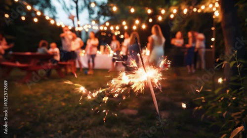A brightly lit sparkler in the foreground, casting a warm glow, with a festive backyard party in the background. 4th july, memorial. independence