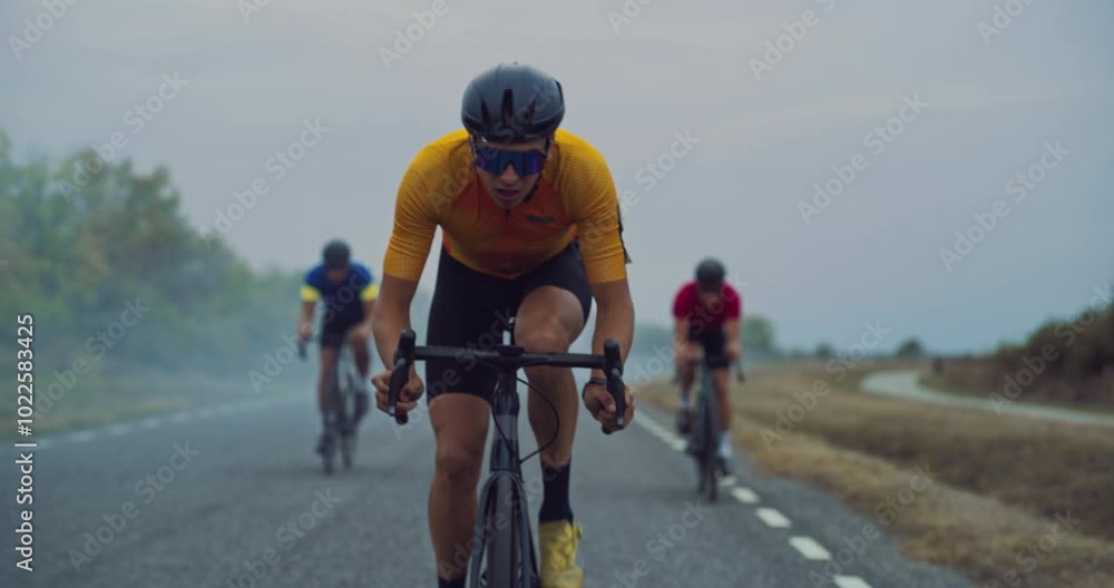Male Cyclist Sets the Pace, Riding Strongly in Front While Two Teammates Follow Behind Side by Side. Athletes Ride Together, Their Focus Intense as They Tackle the Misty Road Ahead Male Cyclist Sets the Pace, Riding Strongly in Front While Two Teammates Follow Behind Side by Side. Athletes Ride Together, Their Focus Intense as They Tackle the Misty Road Ahead