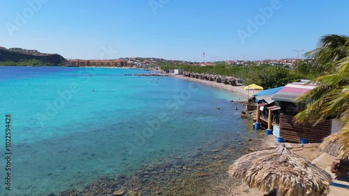 Pristine shoreline at Caracasbaai Beach, Curacao, turquoise waters and lush coastal vegetation under a clear sky with tropical picnic tables on sand photo