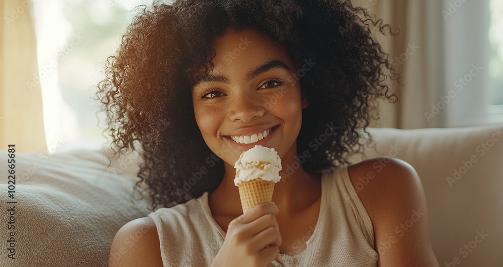 A young Black woman with curly hair smiles while sitting on her couch, eating an ice cream cone. A young Black woman with curly hair smiles while sitting on her couch, eating an ice cream cone.