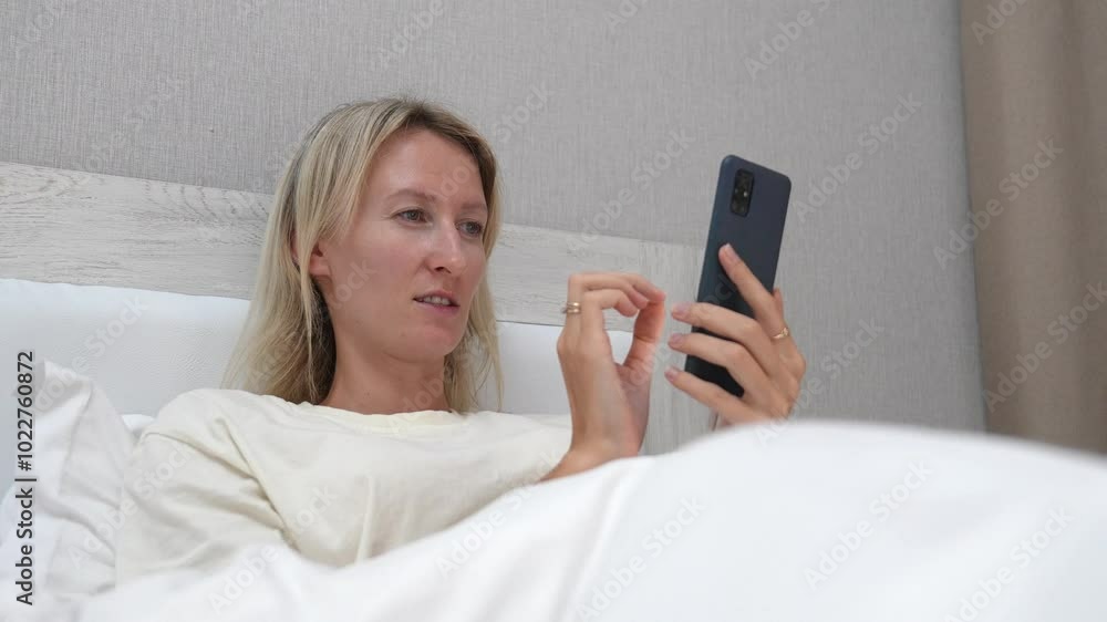 Woman lying in bed with white linens holds smartphone, scrolling and tapping screen while browsing various content online. Relaxation and leisure define scene