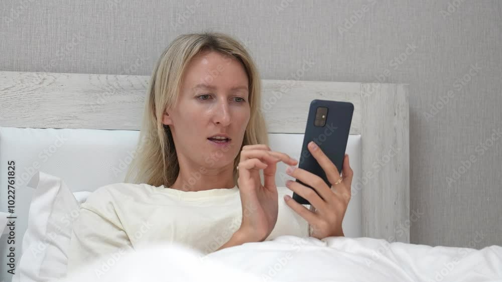Woman lying in bed with white linens holds smartphone, scrolling and tapping screen while browsing various content online. Relaxation and leisure define scene