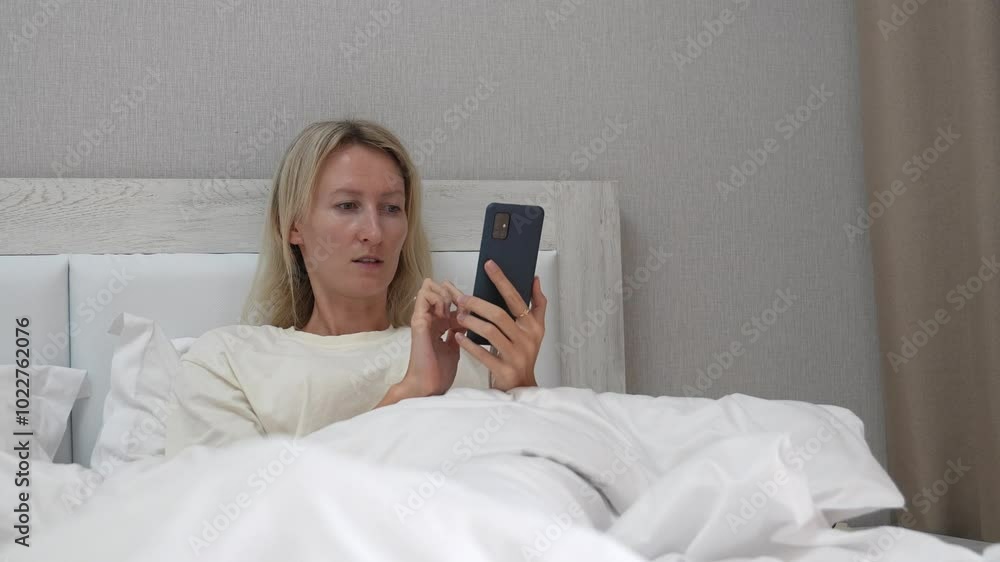Woman lying in bed with white linens holds smartphone, scrolling and tapping screen while browsing various content online. Relaxation and leisure define scene Woman lying in bed with white linens holds smartphone, scrolling and tapping screen while browsing various content online. Relaxation and leisure define scene