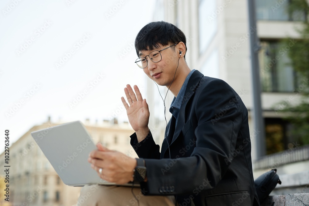 Medium shot of smiling Asian man in headphones looking at laptop screen during online meeting captured saying hello to colleagues while sitting outside in city street, copy space Medium shot of smiling Asian man in headphones looking at laptop screen during online meeting captured saying hello to colleagues while sitting outside in city street, copy space