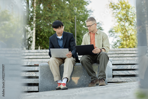 Full shot of male IT programmers using laptops during teamwork on writing code while sitting on concrete bench outside in city enjoying sunny day, copy space