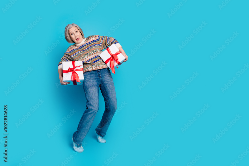 Full length photo of astonished person wear ornament pullover holding two heavy present boxes staring isolated on blue color background Full length photo of astonished person wear ornament pullover holding two heavy present boxes staring isolated on blue color background