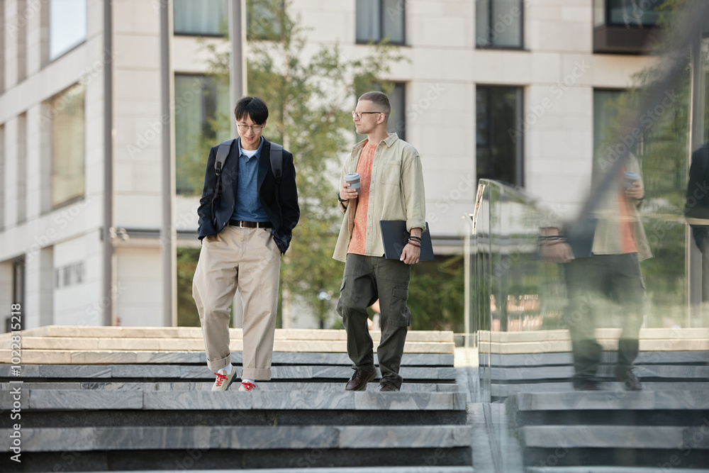 © Seventyfour - Full length shot of two male IT developers in stylish outfits talk business during walking meeting or daily commute to work while descending steps outside in city street, copy space