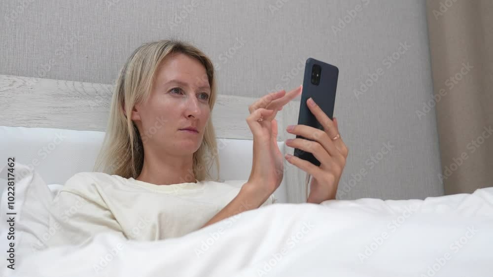 Woman lying in bed with white linens holds smartphone, scrolling and tapping screen while browsing various content online. Relaxation and leisure define scene Woman lying in bed with white linens holds smartphone, scrolling and tapping screen while browsing various content online. Relaxation and leisure define scene