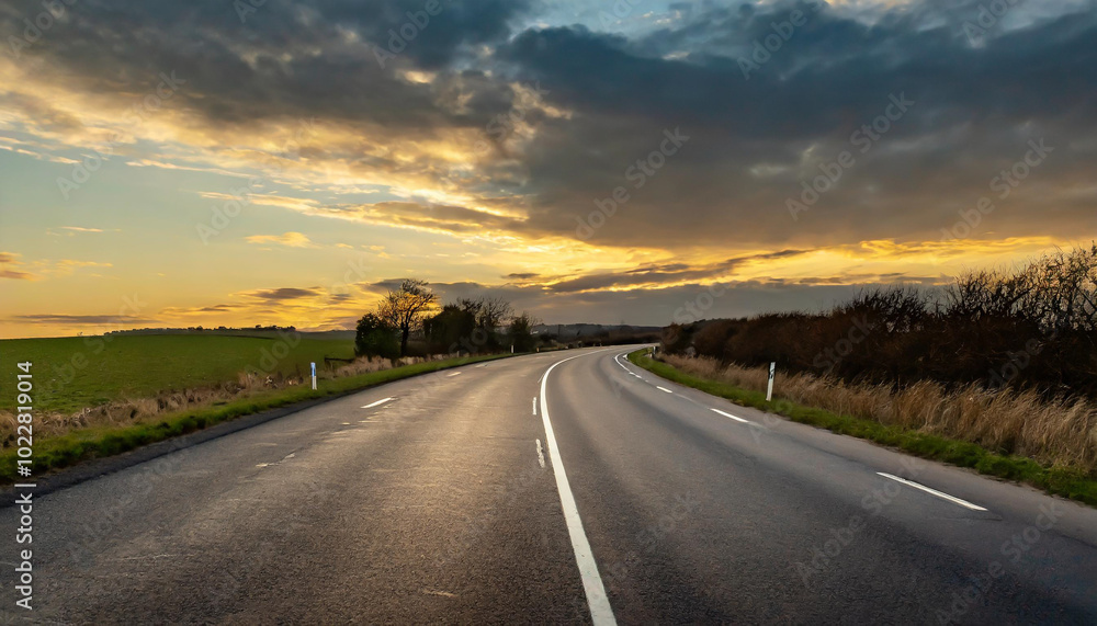Naklejka premium A winding country road at sunset with a vibrant sky and grassy fields in the background