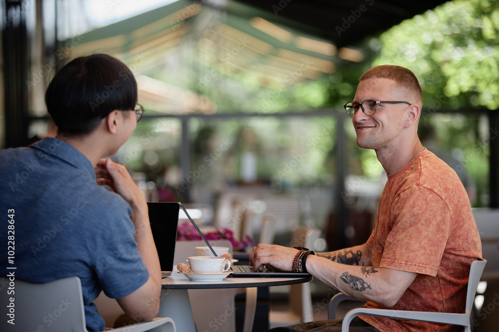 Side view of smiling businessman using laptop sitting at cafe table while enjoying talk and collaborate work over cup of coffee with colleague in outdoor seating area of cafe, copy space Side view of smiling businessman using laptop sitting at cafe table while enjoying talk and collaborate work over cup of coffee with colleague in outdoor seating area of cafe, copy space