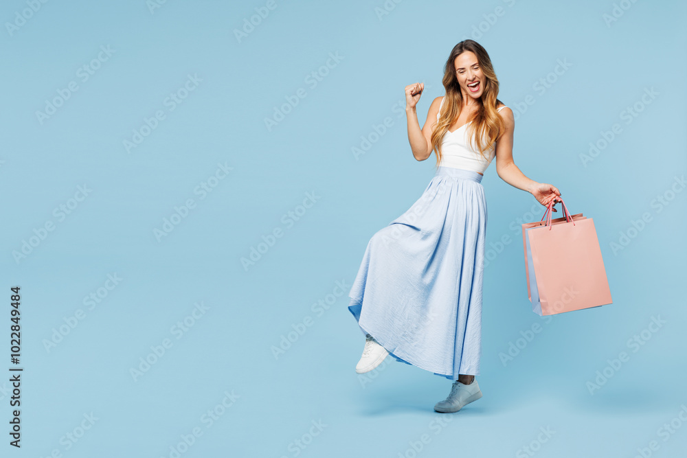 Full body young woman wears casual clothes hold in hand paper package bags after shopping do winner gesture celebrate clench fists isolated on plain blue background. Black Friday sale buy day concept. Full body young woman wears casual clothes hold in hand paper package bags after shopping do winner gesture celebrate clench fists isolated on plain blue background. Black Friday sale buy day concept.