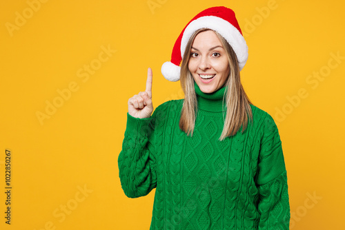 Merry young woman wear green warm cozy sweater Santa hat posing holding index finger up with great new idea isolated on plain yellow background. Happy New Year celebration Christmas holiday concept.