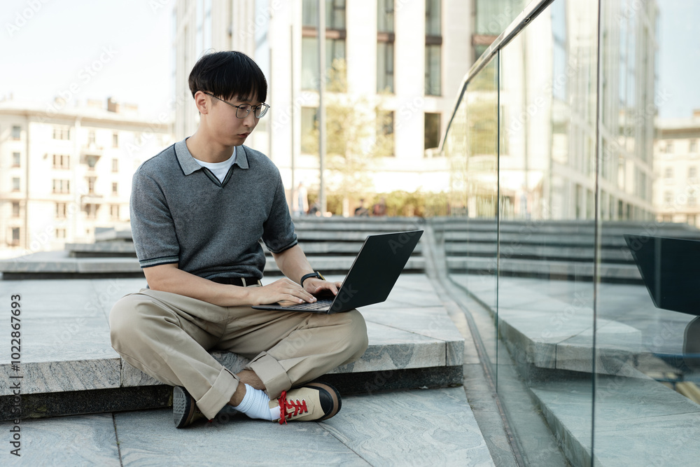 Serious Asian man in stylish gray polo outfit concentrated on using laptop typing on keyboard while sitting on concrete steps outside in city street, copy space Serious Asian man in stylish gray polo outfit concentrated on using laptop typing on keyboard while sitting on concrete steps outside in city street, copy space