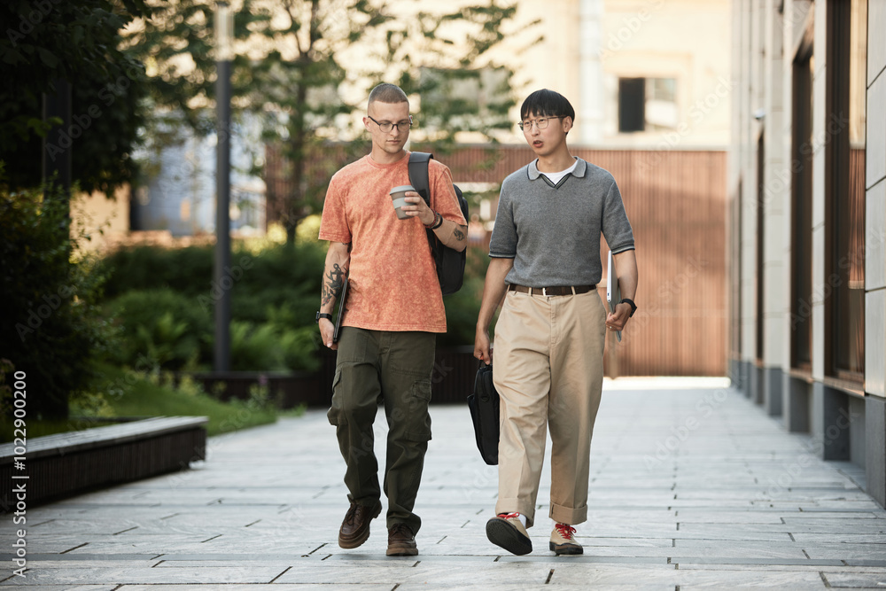 Full length shot of successful male boss and manager chatting holding laptops while walking along business building outside in city street, copy space Full length shot of successful male boss and manager chatting holding laptops while walking along business building outside in city street, copy space