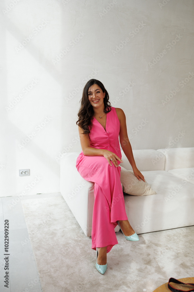 A beautiful tall brunette with spectacular styling stands at the window and smiles. A woman in a pink pantsuit in a vest and trousers on beige sofa. A beautiful tall brunette with spectacular styling stands at the window and smiles. A woman in a pink pantsuit in a vest and trousers on beige sofa.