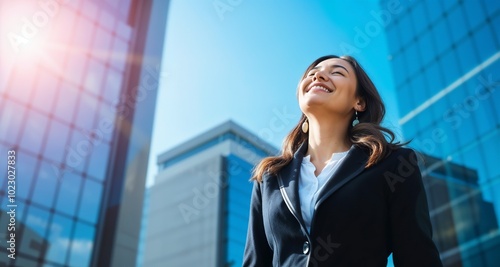 A confident businesswoman stands with closed eyes, basking in the sun amidst towering city buildings, reflecting a sense of peace and accomplishment.