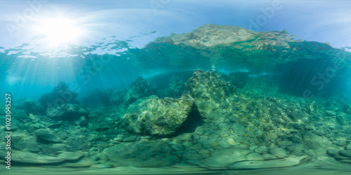 A serene underwater landscape featuring large rocks and clear blue water, captured in a 360-degree panorama. Sunlight filters through the surface, illuminating the rocky seabed and creating a calm atm