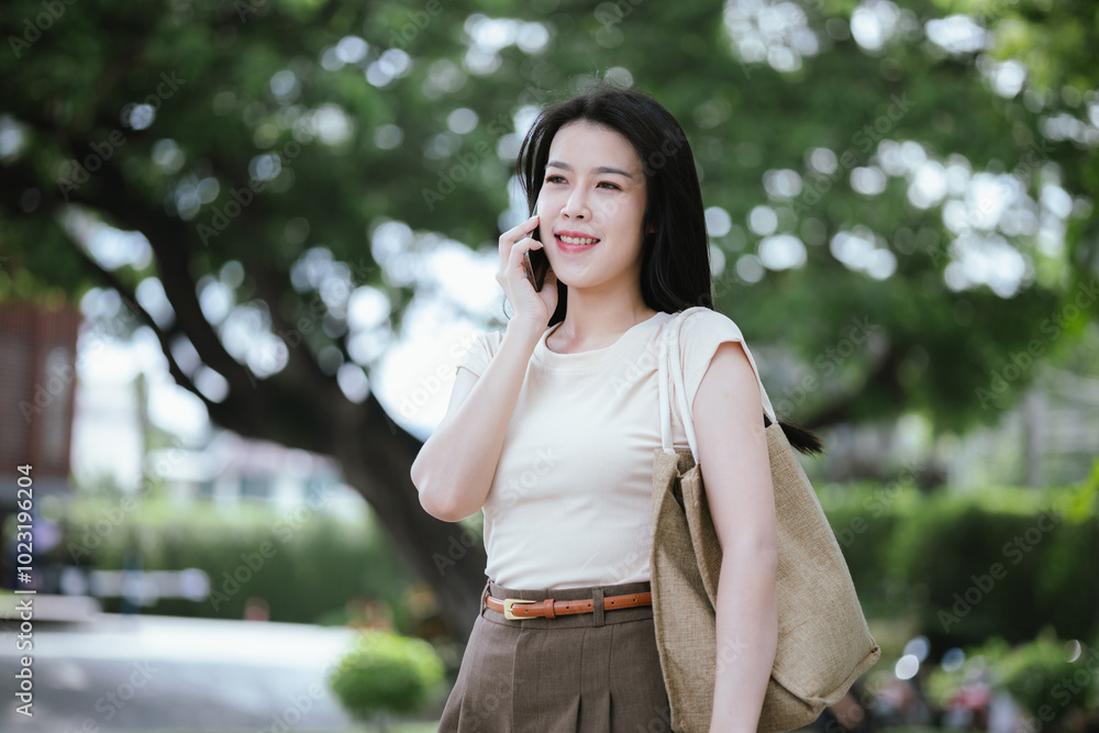 Happy Asian woman sitting on outdoor bench, sipping juice and holding shopping bags. She is relaxing, surfing the web on her smartphone, enjoying a relaxing weekend in the city after a day of shopping