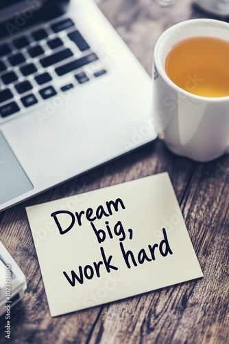 A handwritten note with the quote "Dream big, work hard" placed on a desk next to an open laptop and a cup of tea, in a modern workspace with a motivational vibe.