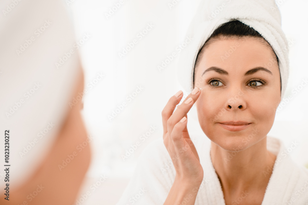 Caucasian middle-aged mature woman in turban after hair washing in bathroom spa looking at the mirror, applying face creme anti-wrinkle rejuvenation effect. Skincare concept Caucasian middle-aged mature woman in turban after hair washing in bathroom spa looking at the mirror, applying face creme anti-wrinkle rejuvenation effect. Skincare concept
