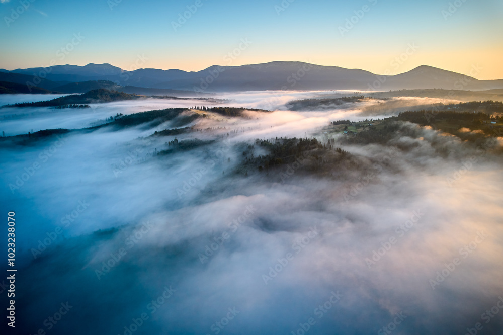 Naklejka premium Stunning aerial view of sunrise over mountainous landscape, with thick fog blanketing valleys and hills. Sun's golden rays pierce through mist, casting serene glow over the entire scene.