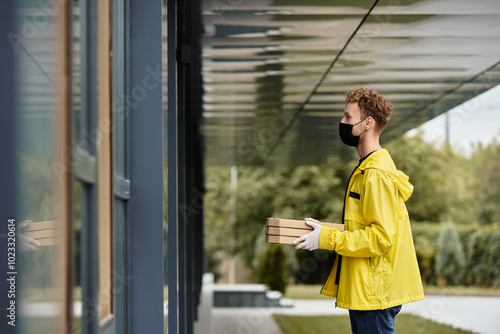 A delivery person in a black mask and yellow jacket arrives at an office building with pizza boxes in hand.