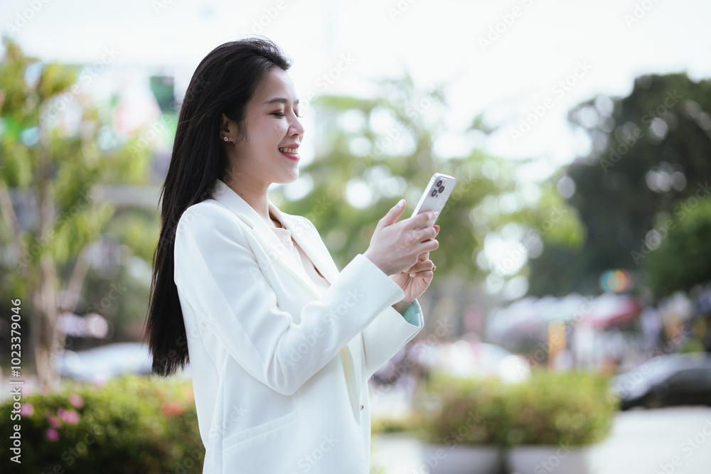 Confident Asian businesswoman in urban suit smiles as she types on her smartphone, balancing a career in digital finance, online e-commerce, and global business opportunities. Confident Asian businesswoman in urban suit smiles as she types on her smartphone, balancing a career in digital finance, online e-commerce, and global business opportunities.