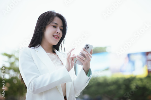 Confident Asian businesswoman in urban suit smiles as she types on her smartphone, balancing a career in digital finance, online e-commerce, and global business opportunities.