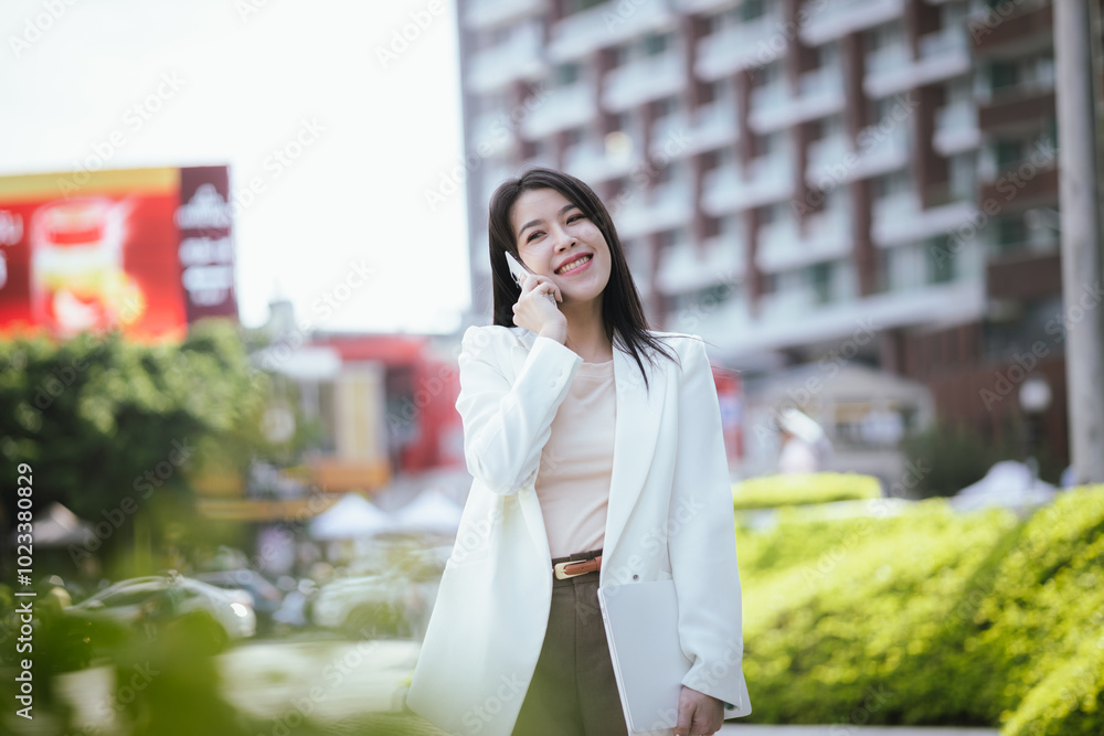 Fototapeta premium Active Asian woman in suit standing outdoors in city holding smartphone and laptop. She represents the future of finance and investment, keeping updated on news and opportunities.