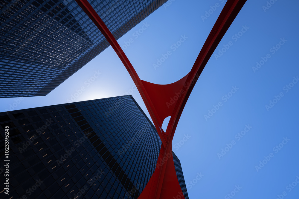 Fototapeta premium Looking up from below, sleek modern skyscrapers are framed by a bold, red steel abstract structure against a clear blue sky. The contrasting elements create a striking, upward perspective.