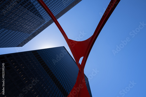 Looking up from below, sleek modern skyscrapers are framed by a bold, red steel abstract structure against a clear blue sky. The contrasting elements create a striking, upward perspective.